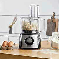 Bosch foodprocessor med deigtilbehør og gjennomsiktig bolle på kjøkkenbord med egg og mel